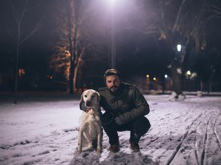A white man in his thirties is squatting next to a blonde labrador retriever. He is wearing a green winter coat, gloves, jeans, and brown shoes. There is snow on the ground, it is nighttime. 